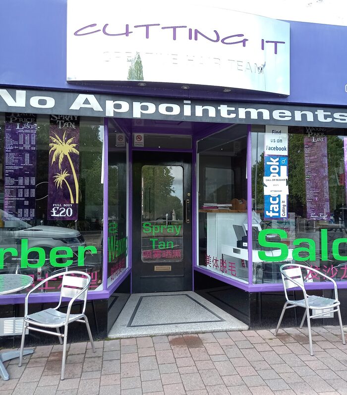 The salon doorway close up, with Barber, Waxing, Spray Tan and Salon lettered on the glass in green and Chinese characters underneath each one, a Spray Tan Full Body £20 banner in the left window and a Find us on Facebook decal in the right
