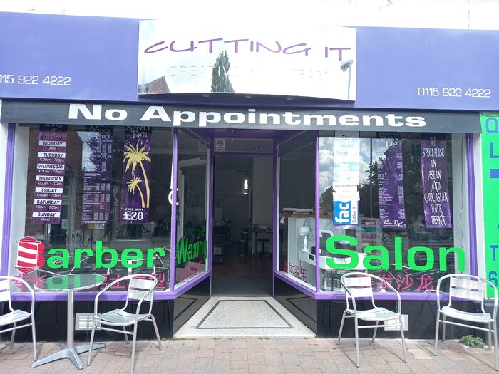 The whole front of the salon on High Road: a purple frontage with a white sign reading Cutting It Creative Hair Team, No Appointments painted in white across the black band above the windows, price lists and an opening hours board in the glass, and two tables with metal chairs on the pavement outside
